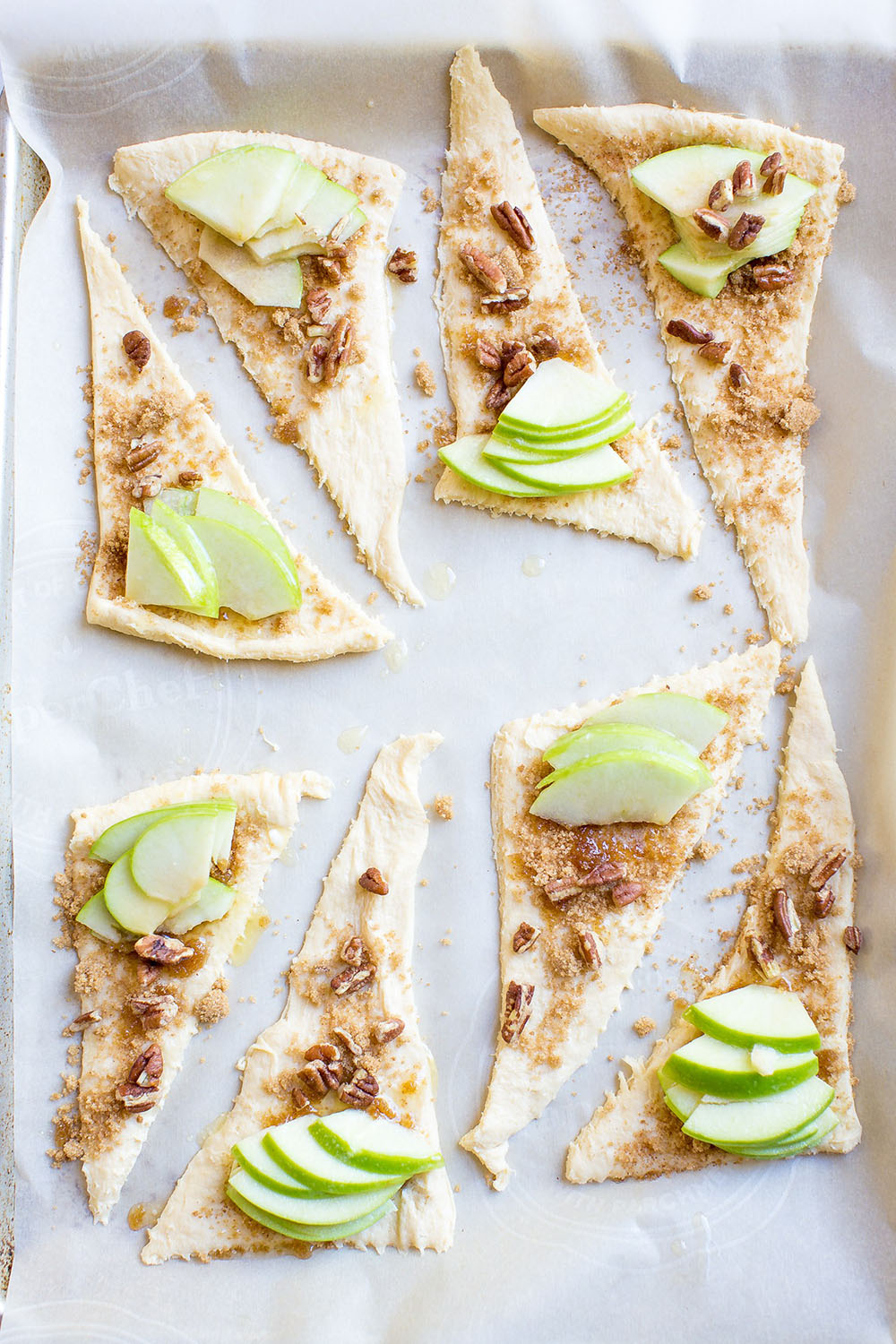 Triangular cuts of crescent dough topped with brown sugar, walnuts and apples on baking try lined with parchment paper.