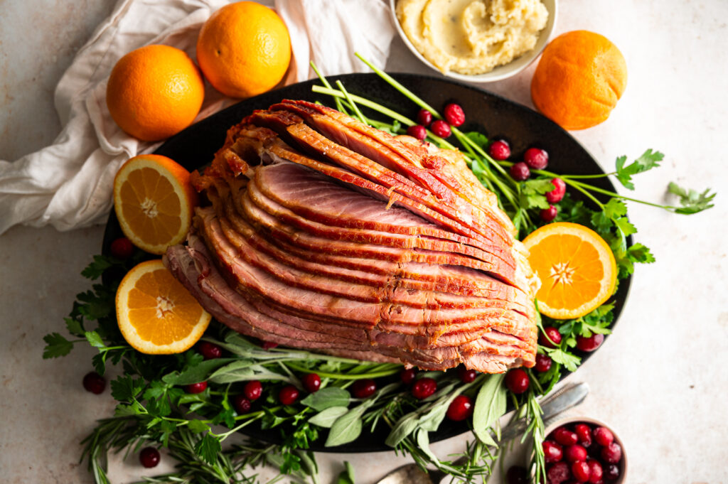 Spiral sliced ham on a plate beneath herbs, surrounded by sliced oranges and cranberries.
