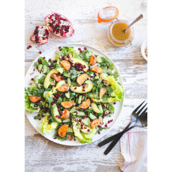 Arugula salad on a plate, topped with avocado, pomegranate, cucumbers and walnuts.