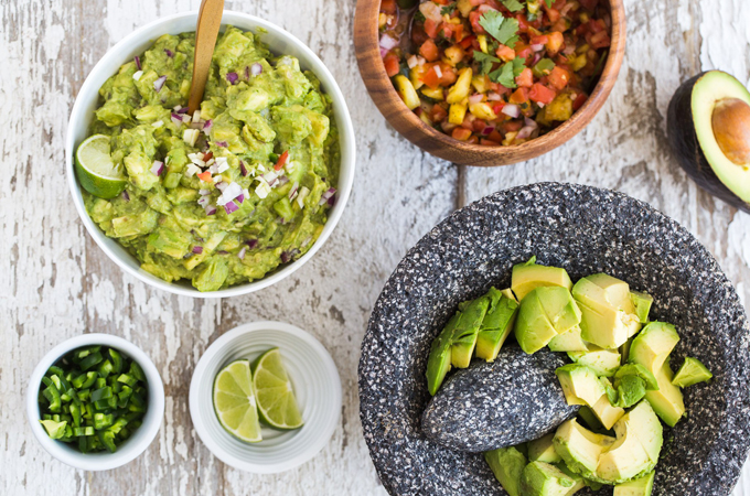 Bowls with guacamole, jalapeños, lime, salsa, and a mortar and pestle with chopped avocados.