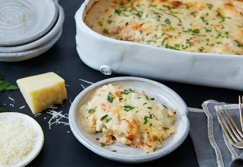 Casserole dish with white lasagna bechamel sauce and smoked salmon next to a plate with a piece of the lasagna.
