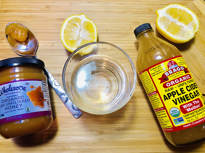 Cutting board with Wholesome raw unfiltered honey, Bragg raw unfiltered apple cider vinegar, lemons and a small glass dish filled with a clear liquid.
