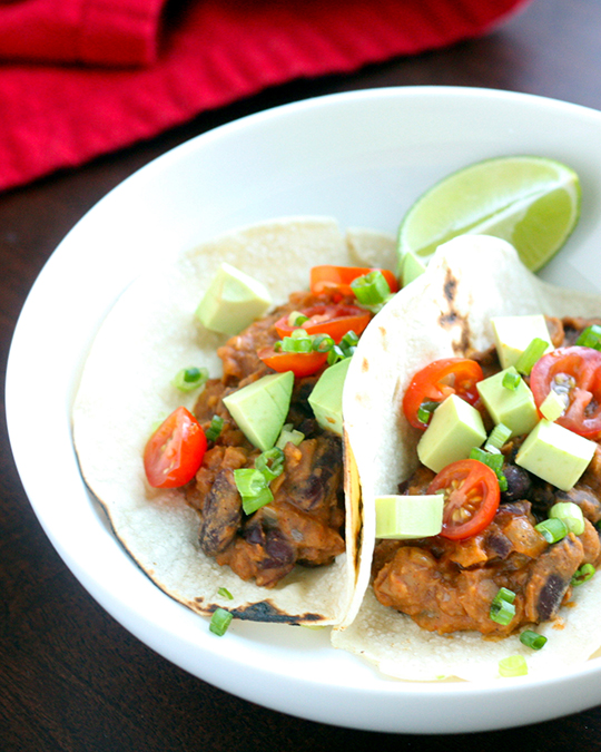Tacos filled with a mix of beans, topped with fresh avocado and diced tomatoes, and served on a plate with a wedge of lime.