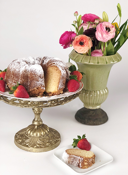 Cake stand with a ricotta bunt cake topped with powdered sugar, surrounded by strawberries, next to a vase with flowers.