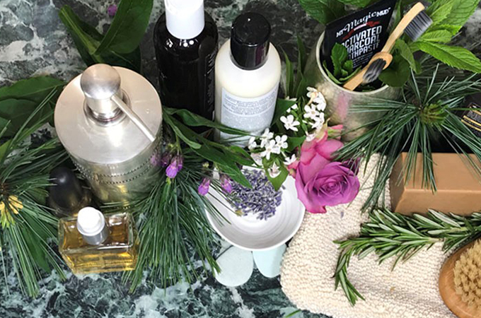 Counter with fresh herbs and leaves surrounding health and beauty bottles, perfumes and soaps.
