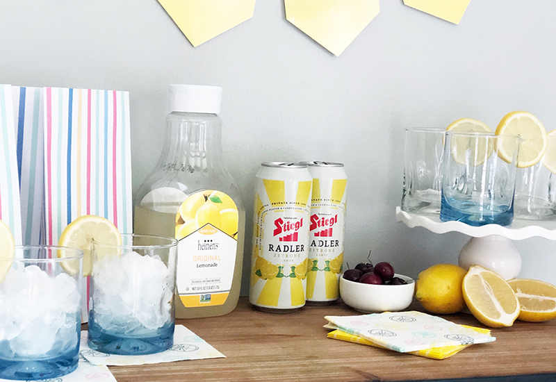 Table with party setting and glasses with ice, container of Heinen's Original Lemonade, cherries, lemons, and Stiegl Lemon Radler cans.