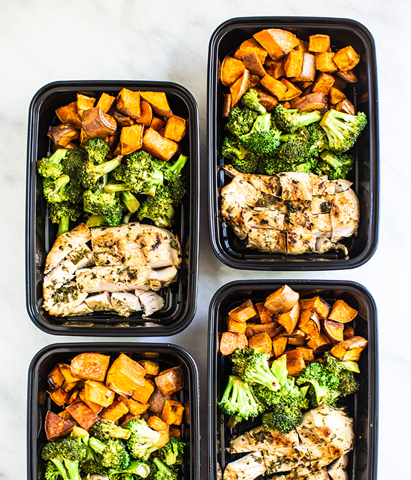 Four black meal prep containers filled with sweet potatoes, broccoli and chicken.
