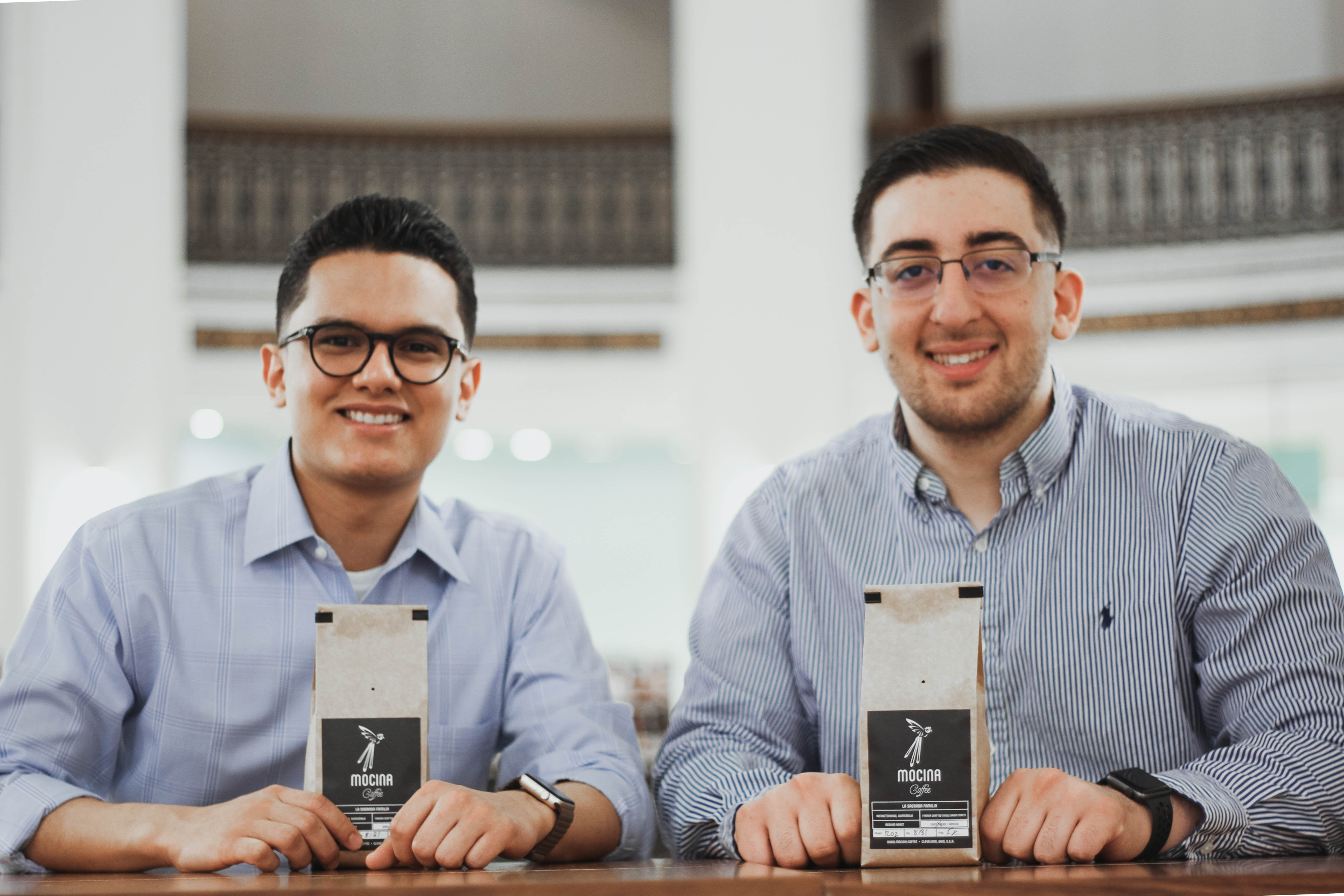 Two men sitting at a table with bags of Mocina coffee beans.