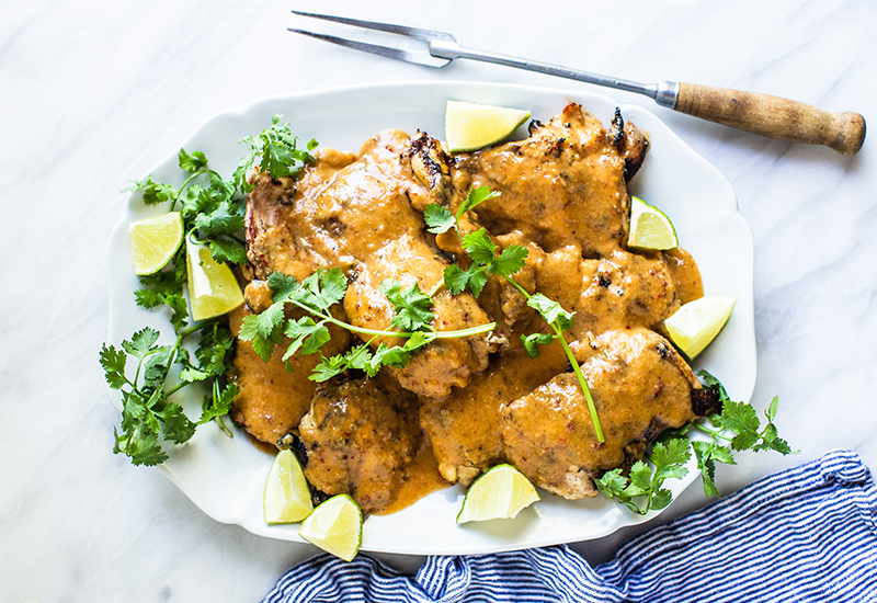 Chicken on a plate in a creamy sauce with sprigs of cilantro and quartered limes.