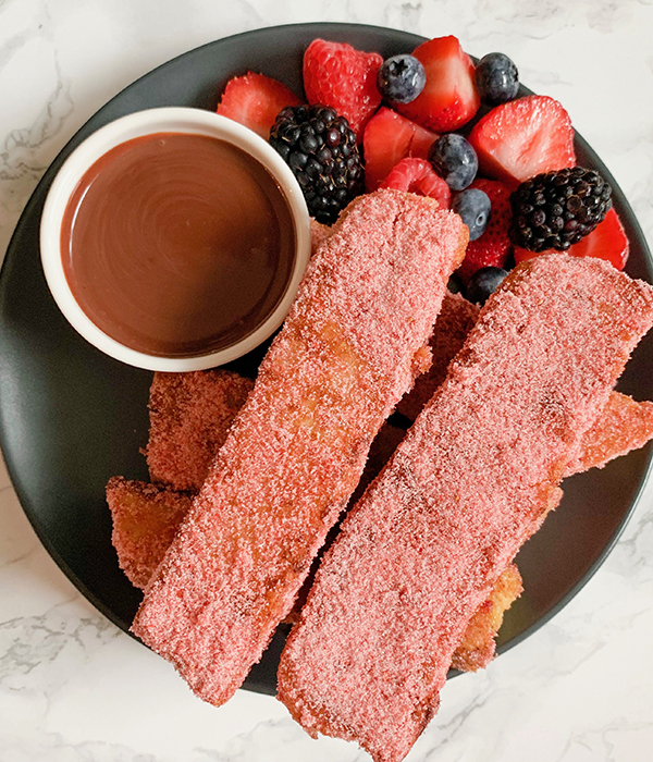 Plate with strawberry French toast sticks coated in freeze-dried strawberries next to chocolate dipping sauce and mixed berries.