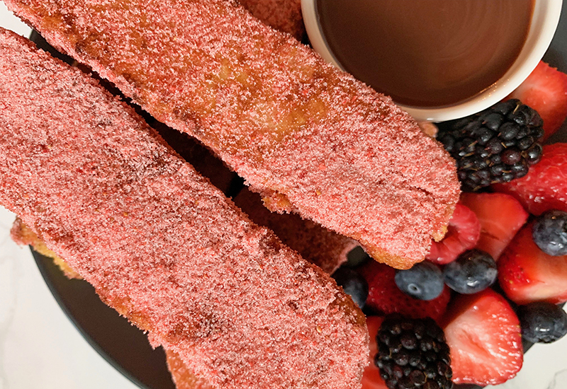 Plate with strawberry French toast sticks coated in freeze-dried strawberries next to chocolate dipping sauce and mixed berries.