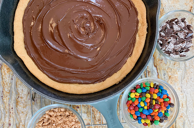 Sugar cookie in a large cast iron skillet topped with melted chocolate next to bowls with toffee bits, nonpareils and mini M&Ms.
