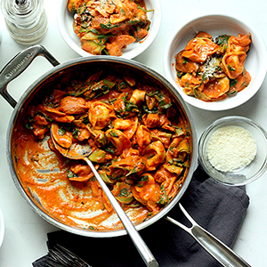 Skillet with tri-colored tortellini in a spinach tomato sauce next to parmesan Reggiano cheese and bowls of pasta.