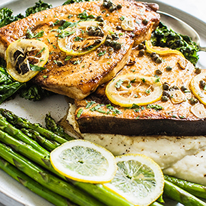 Plated with swordfish fillets topped with sliced lemons and cappers over mashed potatoes with a side of asparagus.