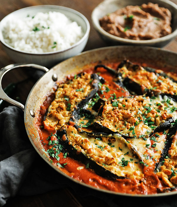 Skilled with a tomato sauce and chiles stuffed with chicken and cheese next to bowls of white rice and refried beans.