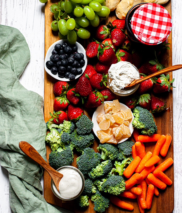 Cutting board with broccoli florets, carrots, ranch dipping sauce, strawberries, mandarin oranges, blueberries and grapes and a fruit dip.