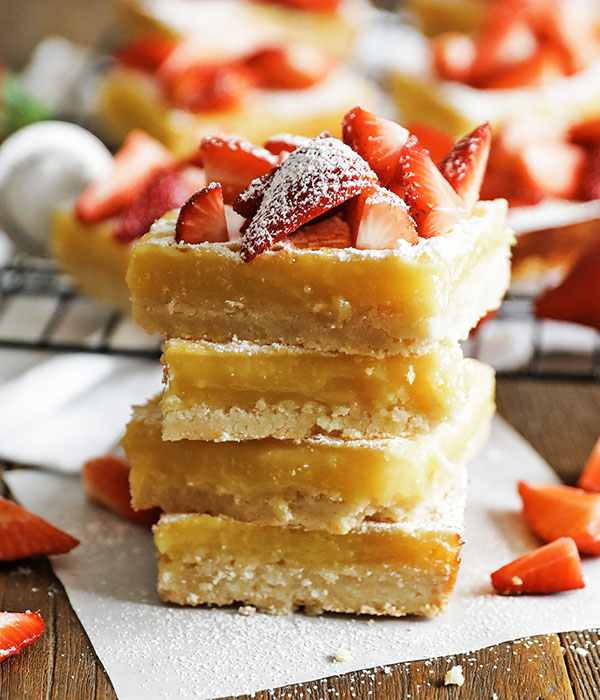Stack of square-cut lemon bars with a graham cracker crust topped with sliced strawberries and dusted with powdered sugar.