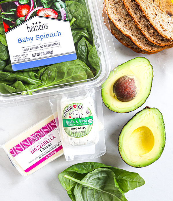 Counter with packaged baby spinach, a halved avocado, block of mozzarella cheese, package of goat cheese, and slices of bread.