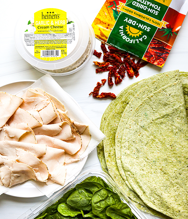 Ingredients on a kitchen counter including spinach wraps, sliced turkey breast, spinach, sundried tomatoes and cream cheese.