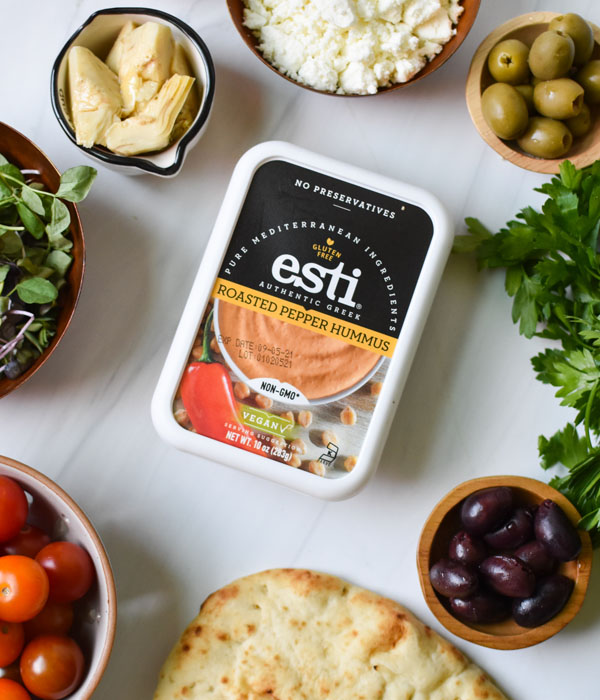 Esti roasted red pepper hummus on a kitchen counter surrounded by feta cheese, flatbread, tomatoes, artichoke hearts, and olives.
