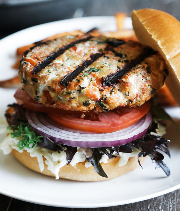 Salmon burger on a plate topped with coleslaw, spring mix, red onion and tomato.