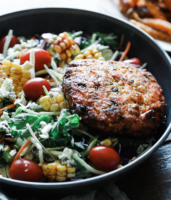 A bowl of salad with cheese, corn, and tomatoes topped with a fresh salmon burger.