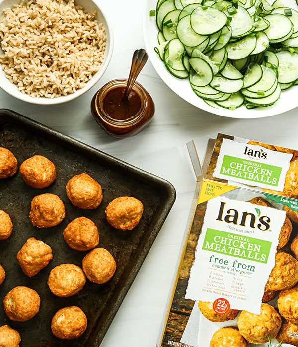 Baking dish with meatballs next to a jar of BBQ sauce, bowls of sliced cucumbers and brown rice, and box of Ian's Chicken Meatballs.