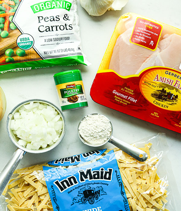 Counter with egg noodles, chicken breast, onions, flour, poultry seasoning, and peas and carrots.