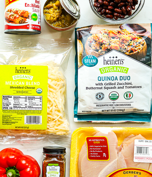 Ingredients arranged on a counter, including chicken breasts, dry seasonings, olive oil, onion, red bell pepper, cloves, quinoa with veggies, enchilada sauce, black beans, corn, shredded cheese, and cilantro.