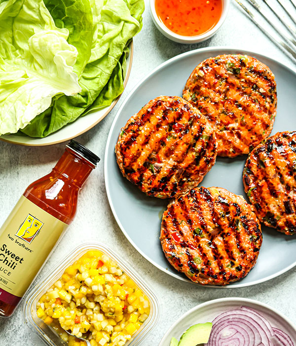 Overhead view of a plate of salmon burgers next to butter lettuce, pineapple mango salsa and Two Brother's sweet chili sauce.