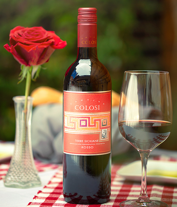 Table with a red and white checkered place mate with a bottle of Colosi Rosso wine next to a wine glass.