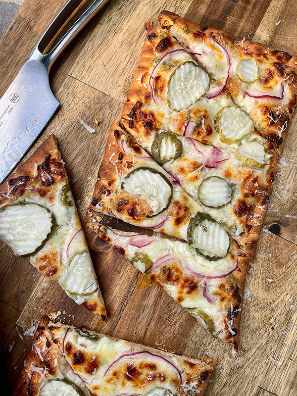 Rectangular flatbread pizza topped with mozzarella cheese, pickles and red onion on a cutting board.