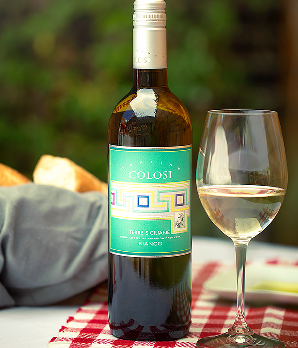 Table with a red and white checkered place mate with a bottle of Colosi Bianco wine next to a wine glass.