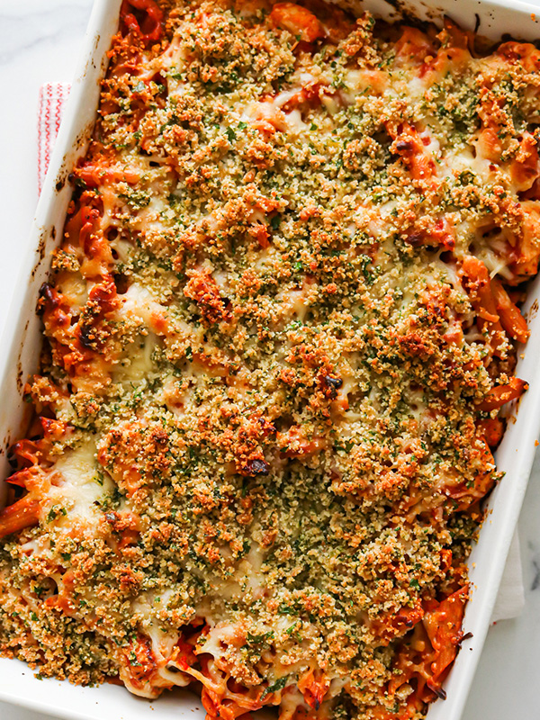 White baking dish with penne pasta and marinara sauce topped with melted mozzarella cheese and panko bread crumbs.