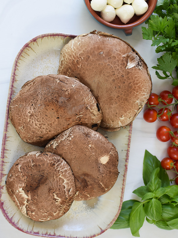 Portobella mushrooms on a plate next to basil, parsley, tomatoes and mozzarella balls.