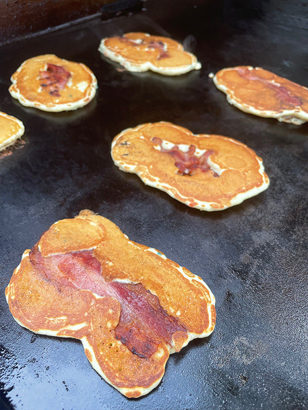 Pancakes with a strip of bacon on a flat-top griddle.