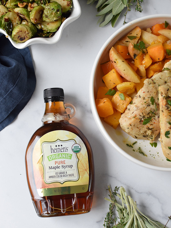 Apple and butternut squash served with chicken breasts on a plate next to maple syrup and brussels sprouts.