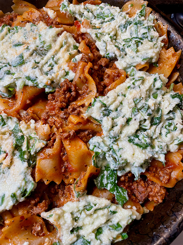 Skillet with lasagna noodles, tomato sauce and ground beef, topped with dollops of ricotta and spinach.