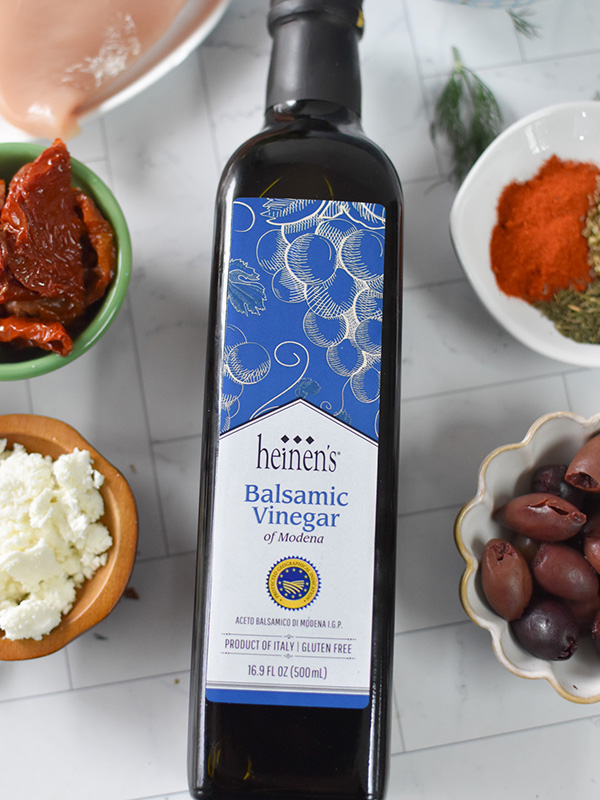 Bottle of Heinen's balsamic vinegar surrounded by feta cheese, olives, sundried tomatoes, and spices.