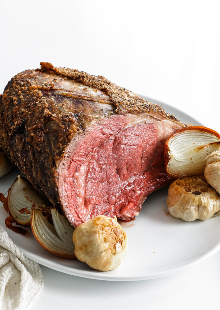Standing rib roast served on a platter with roasted halved onions and whole garlic bulbs.