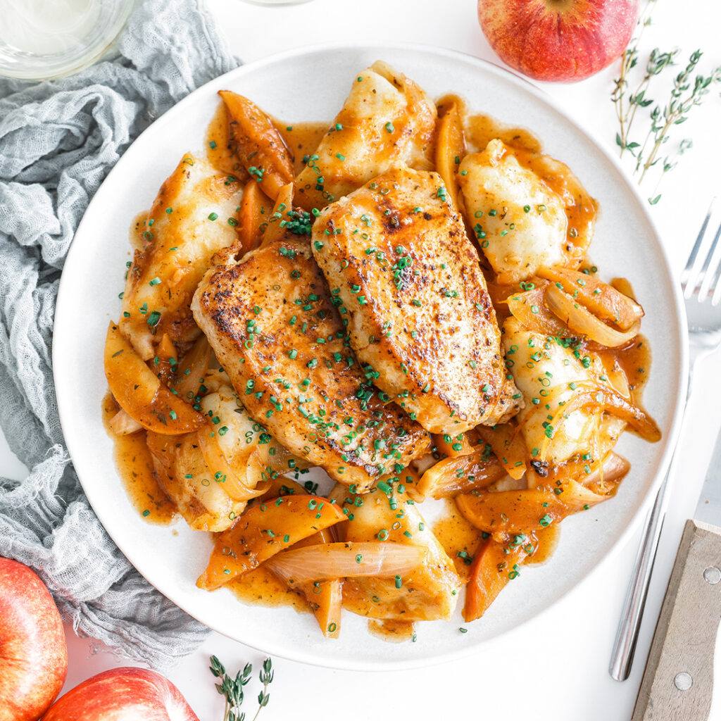 Pan-seared pork chops served over a plate of apples and pierogis with onions.