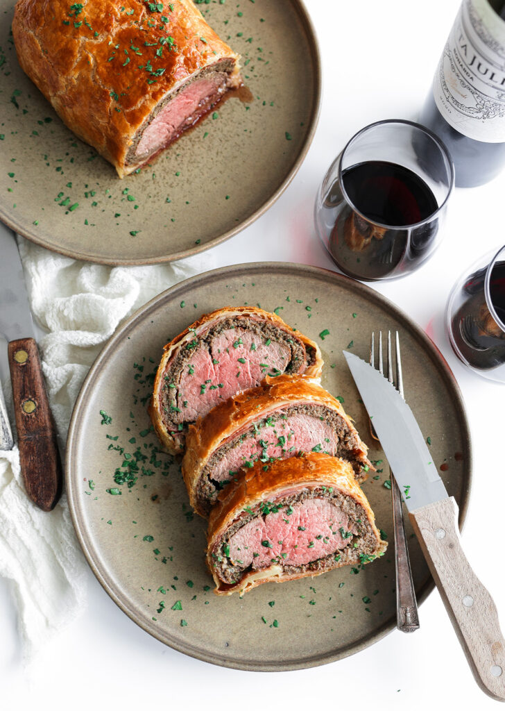 Slices of beef wellington served on a plate with a glass of wine.
