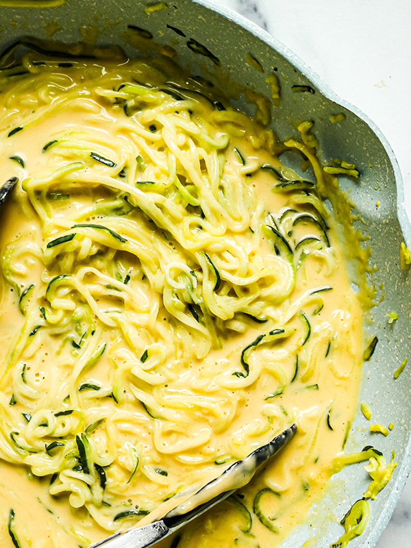 Overhead view of zucchini spirals being cooked in a cheddar cheese sauce in a skillet.