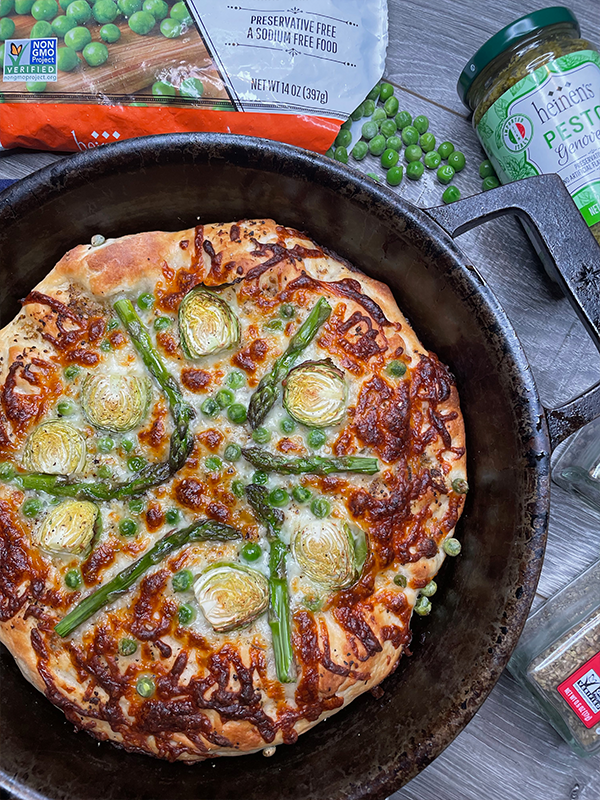 Pizza in a cast iron skillet topped with pesto, cheese, peas, brussels sprouts, and asparagus.