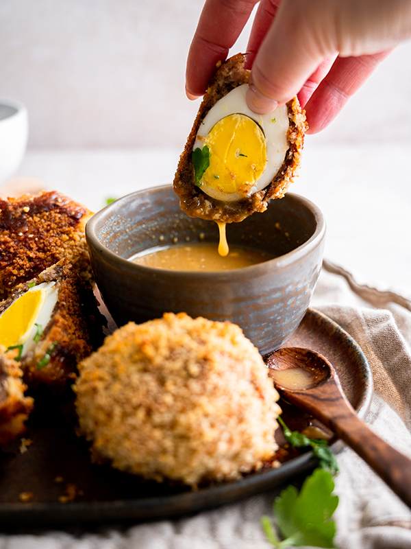 Halved baked scotch egg being dipped into a maple mustard sauce.