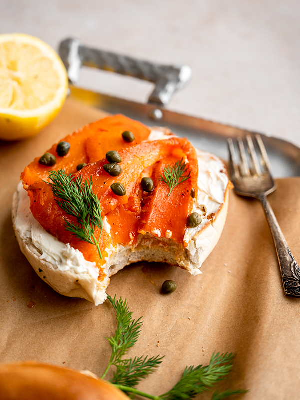 Bagel with cream cheese topped with carrot lox and capers on a cutting board.