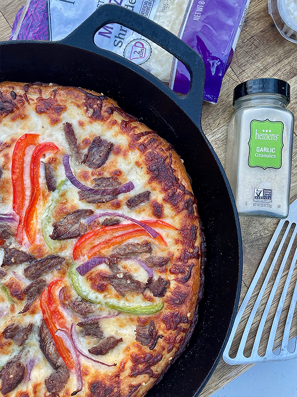 Pizza in a cast iron skillet, topped with steak, bell pepper and onion slices next to shredded mozzarella cheese and garlic powder.