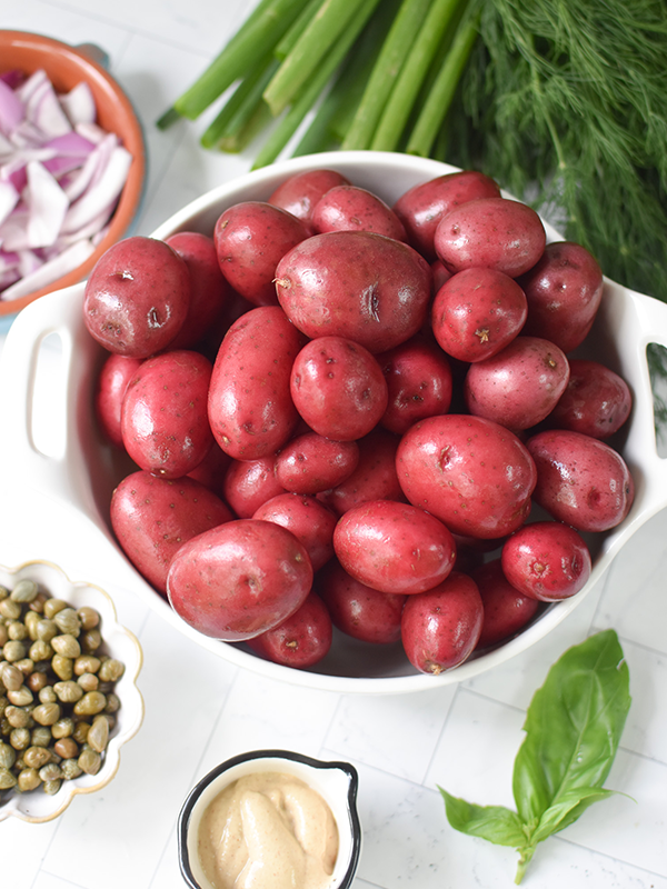 Whole red potatoes in a pot surrounded by dill, celery, red onion, and basil.
