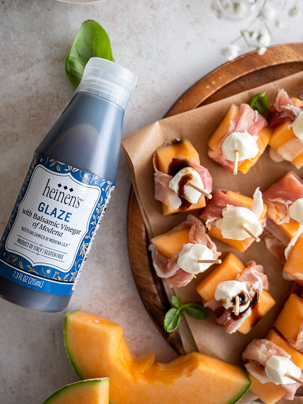 Melon, prosciutto, and mozzarella cheese bites topped with balsamic vinegar glaze, next to a bottle of Heinen's balsamic vinegar glaze.