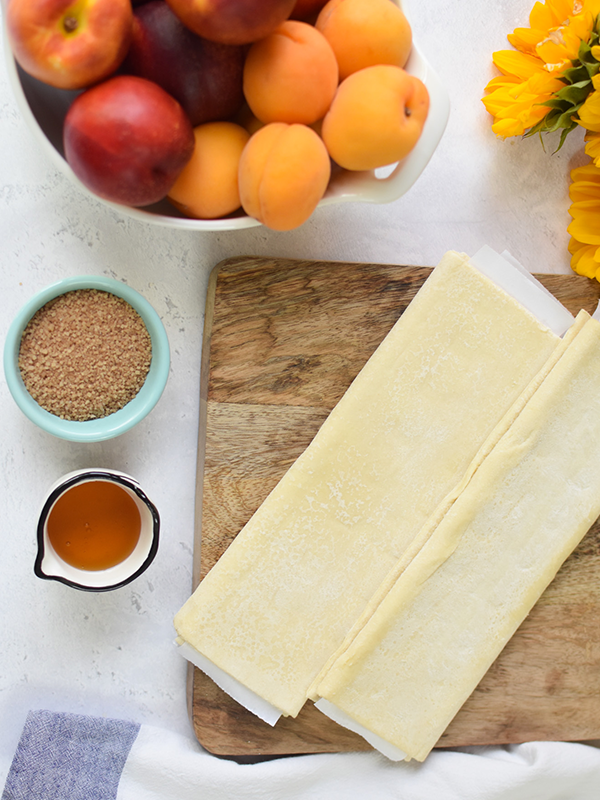 Pastry sheets on a cutting board next to spices and a bowl of stone fruit.